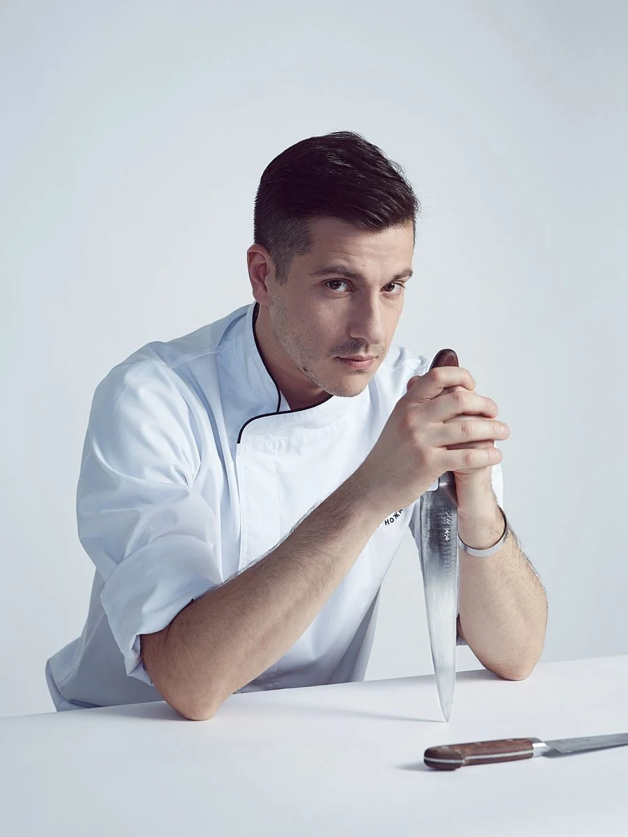 Man in a white chef jacket holding a kitchen knife at a table.