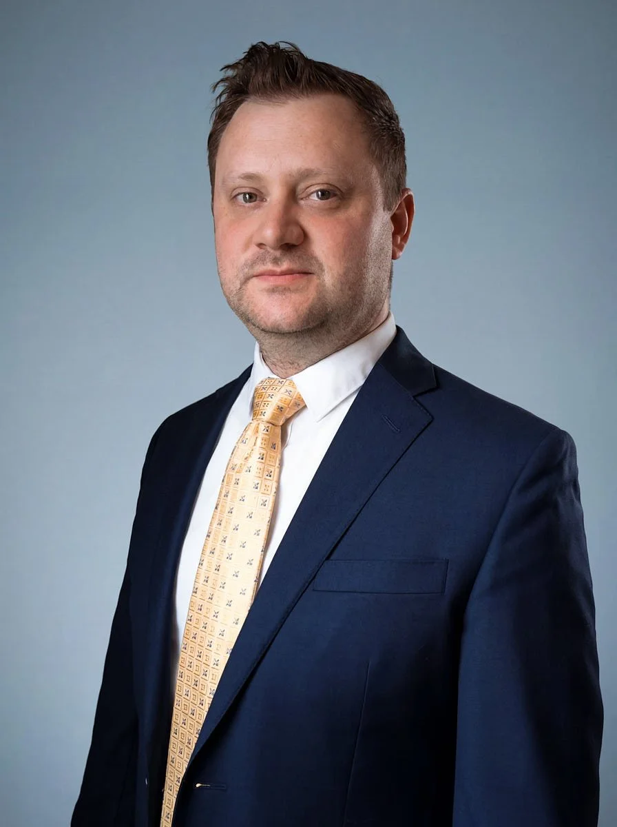 Person in a navy suit and yellow patterned tie against a blue background.