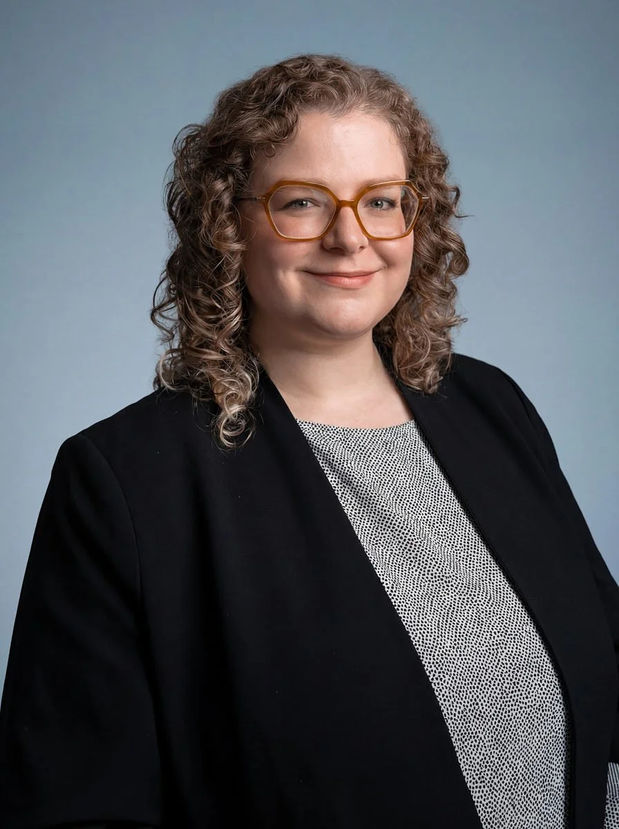 Person with curly hair and glasses wearing a black jacket and gray top.