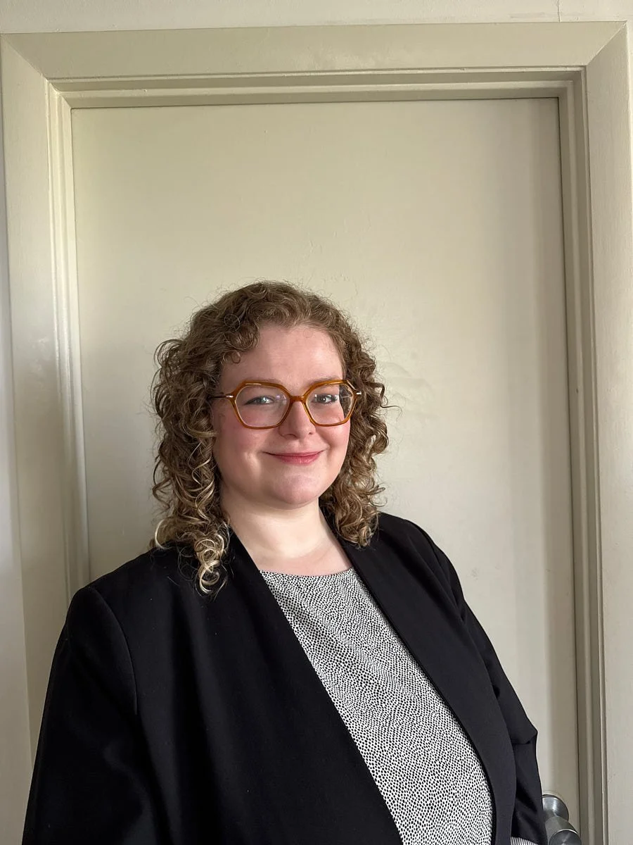Person with curly hair and glasses wearing a black jacket, photographed indoors.